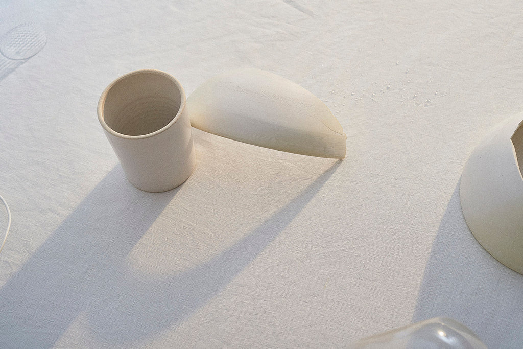 A white, handmade ceramic bowl leans against a white ceramic cup on a white linen tablecloth. 